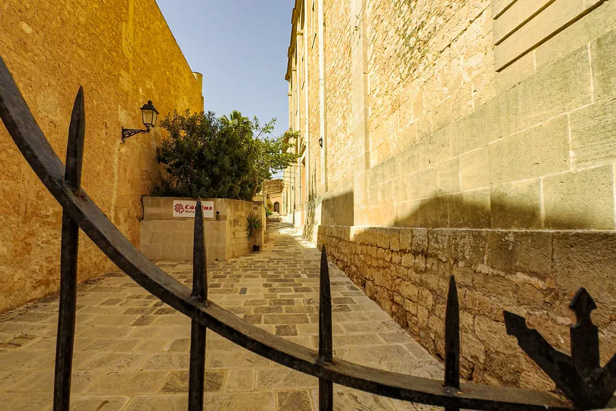 Schmale ansteigende Gasse entlang der massiven Sandsteinmauer der Kirche von Ses Salines, mit Geländer und Straßenlaterne.