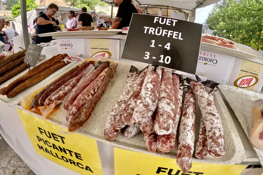 Auslage mit Fuet und luftgetrockneten Würsten auf einer weißen Theke, dahinter ein Schild mit deutscher Aufschrift und Preisen.
