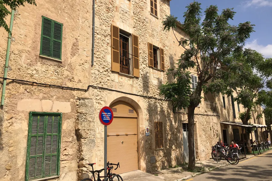 Platz in Sineu mit Sandsteinhäusern, grünen Fensterläden, einem Baum in der Mitte und abgestellten Fahrrädern, im Hintergrund Arkaden.
