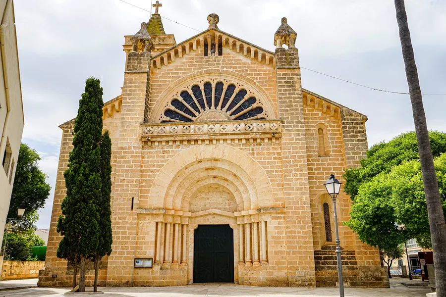 Die Kirche Sant Miquel in Son Carrió aus hellem Sandstein mit Rundbogenportal, halbrunder Fächerrosette und zwei kleinen Türmchen, davor eine Zypresse.
