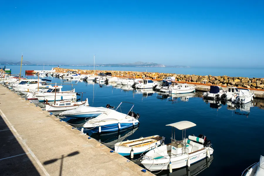 Kleiner Hafen von Son Serra de Marina mit Reihen weißer Motorboote am Steg, dahinter die Steinmole und die Bucht von Alcúdia.