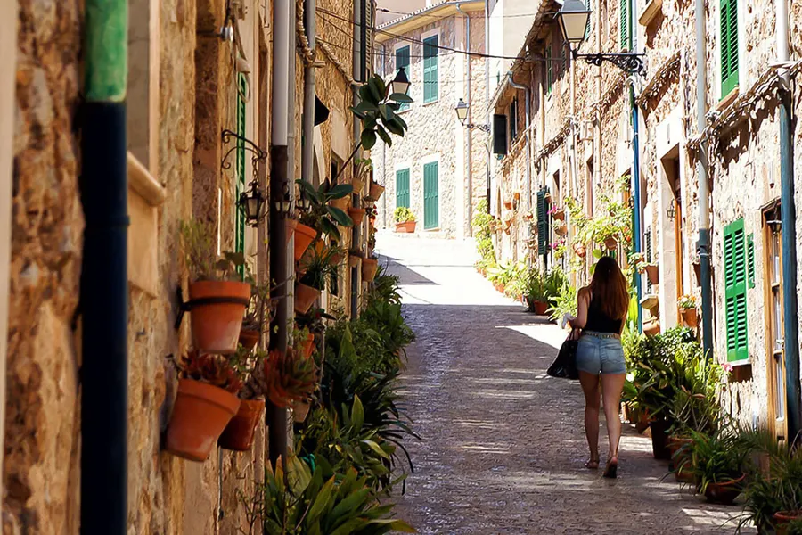 Enge Gasse in Valldemossa mit Natursteinhäusern, grünen Fensterläden und Reihen von Tontöpfen an den Wänden, eine Passantin geht hinauf.