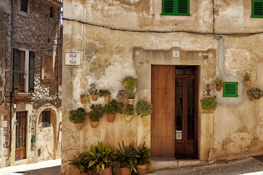 Hausfassade in Valldemossa mit hölzerner Tür, grünen Fensterläden, dem Straßenschild Calle de la Plaza und Topfpflanzen davor.